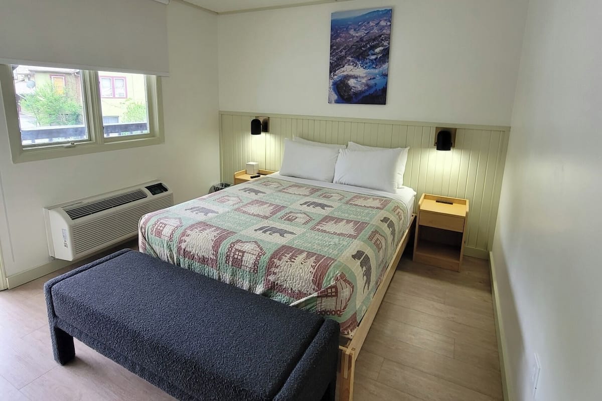 Un dormitorio con una cama, dos mesitas de noche, un banco, un radiador y una ventana.