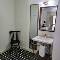 Un baño con piso de baldosas hexagonales blanco y negro, un lavamanos blanco con detalles dorados, una silla negra y un espejo grande.