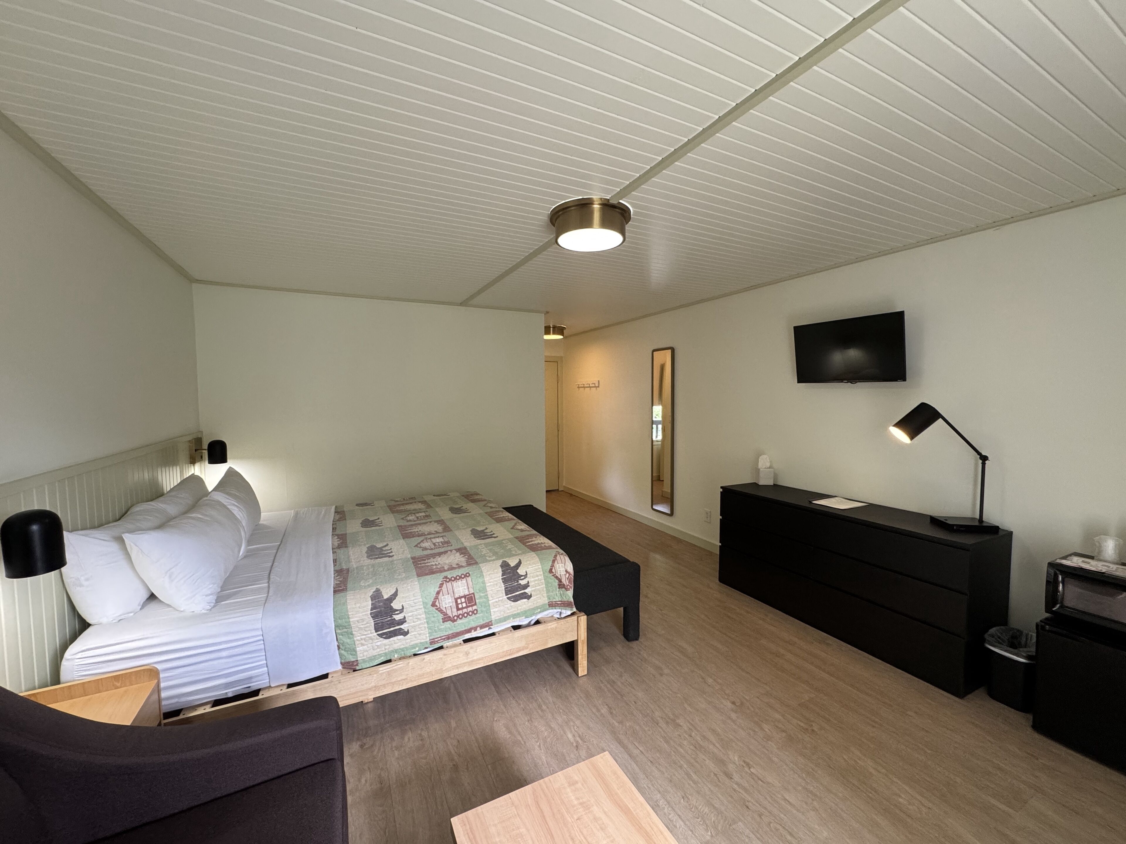 Et soveværelse med seng, natbord, kommode, spejl og fjernsyn på væggen.