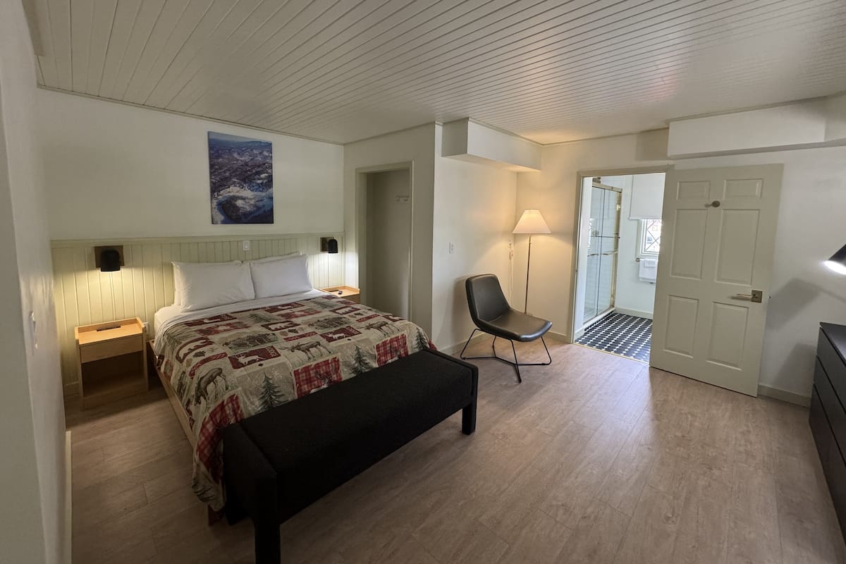 Un dormitorio con cama, mesitas de noche, una silla, una lámpara y una puerta que da a otra habitación.