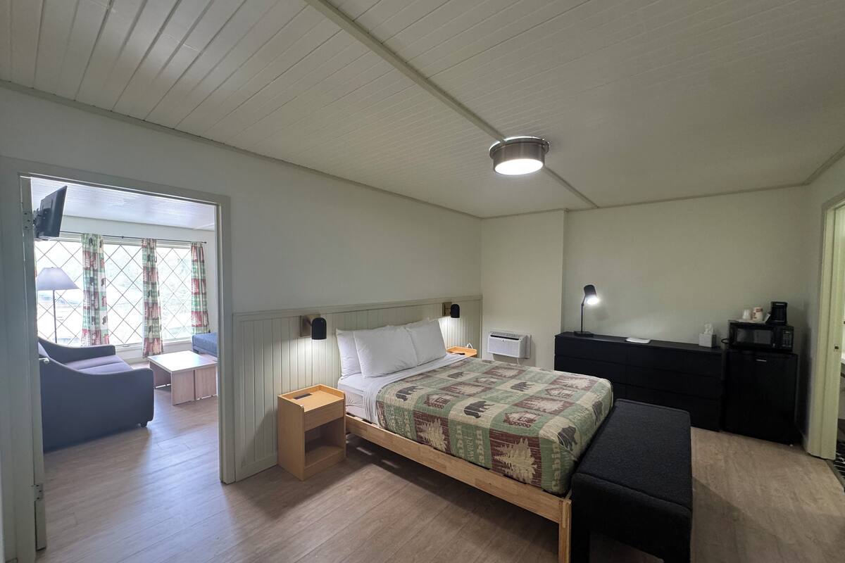 Un dormitorio con cama, mesita de noche, lámpara y un escritorio con silla. Hay una ventana con cortinas y vistas al exterior.