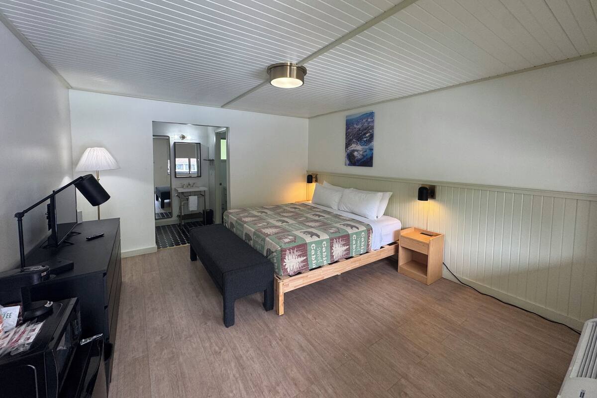 Un dormitorio con cama, mesita de noche, lámpara, escritorio y un pequeño banco.