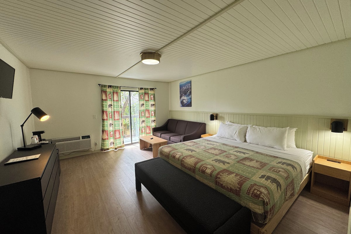 Un dormitorio con cama, una banca, un escritorio con lámpara, un sofá y una ventana con cortinas.