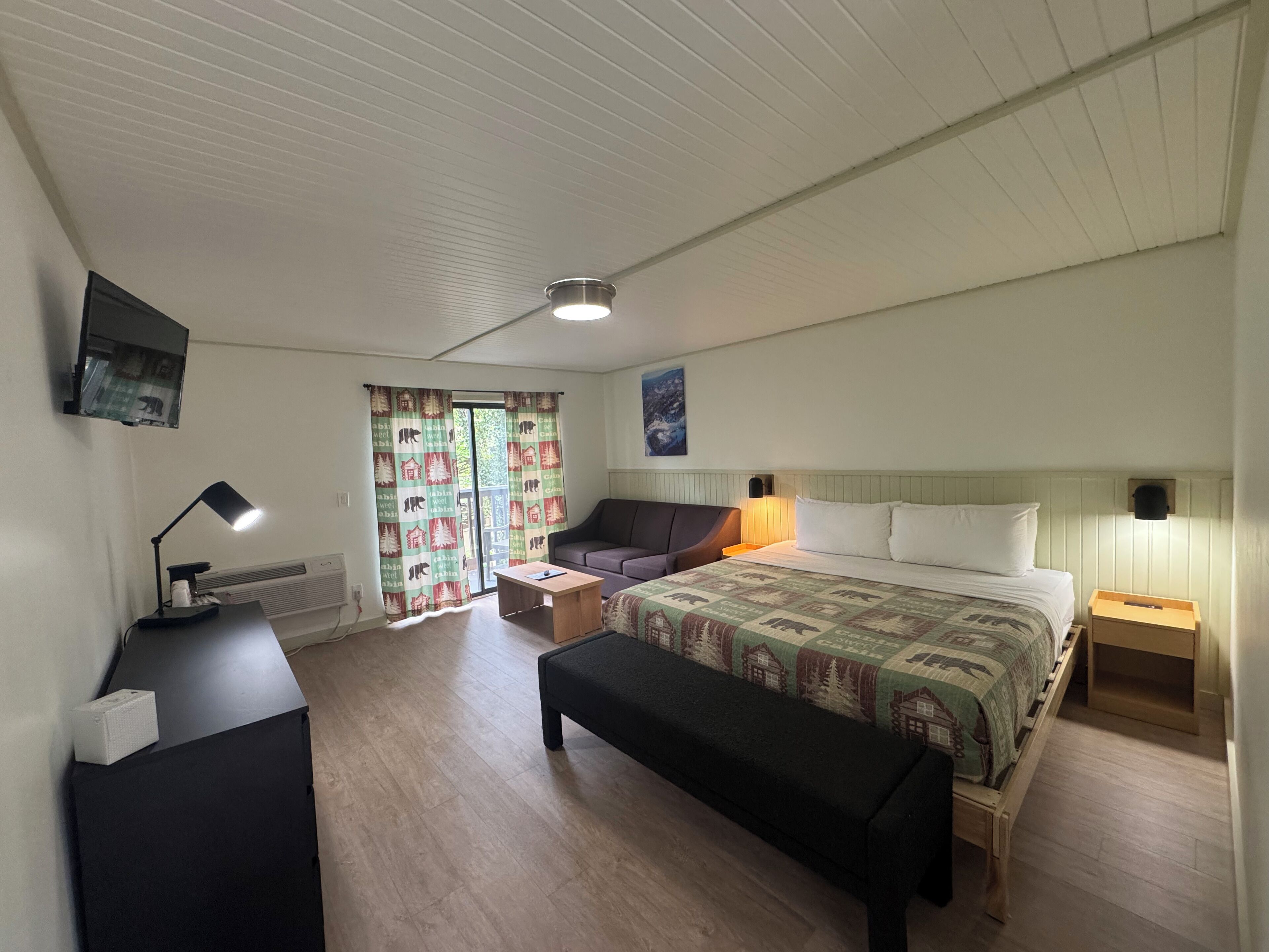 A bedroom with a bed, a desk, a chair, a bench, a TV, and a window with curtains.