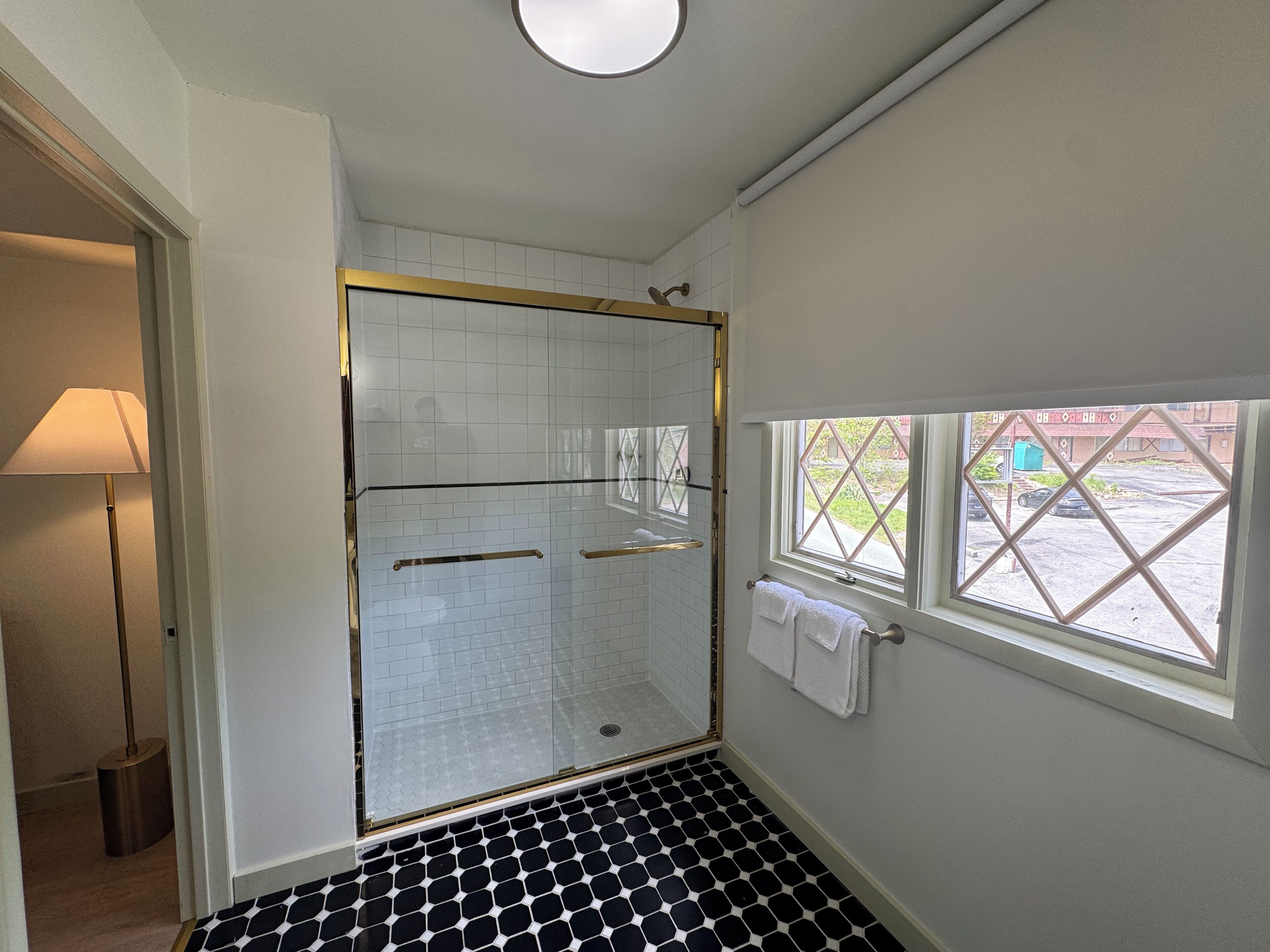 A bathroom with a glass shower enclosure, a black and white tiled floor, a window with a view of the outside, and a towel rack with a white towel.