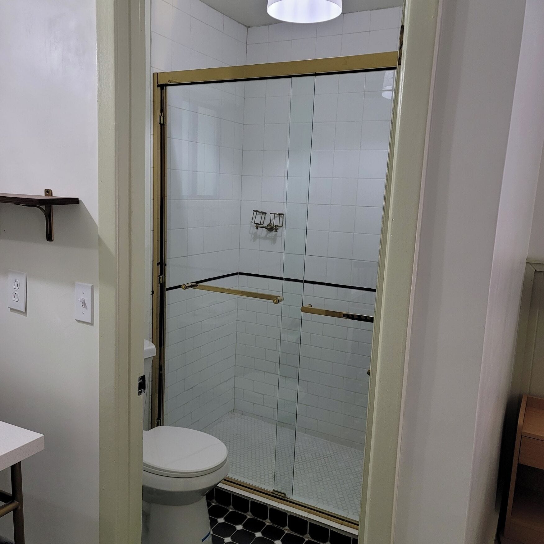 Un baño con ducha de vidrio, paredes cubiertas de azulejos blancos, inodoro blanco y una pequeña estantería.