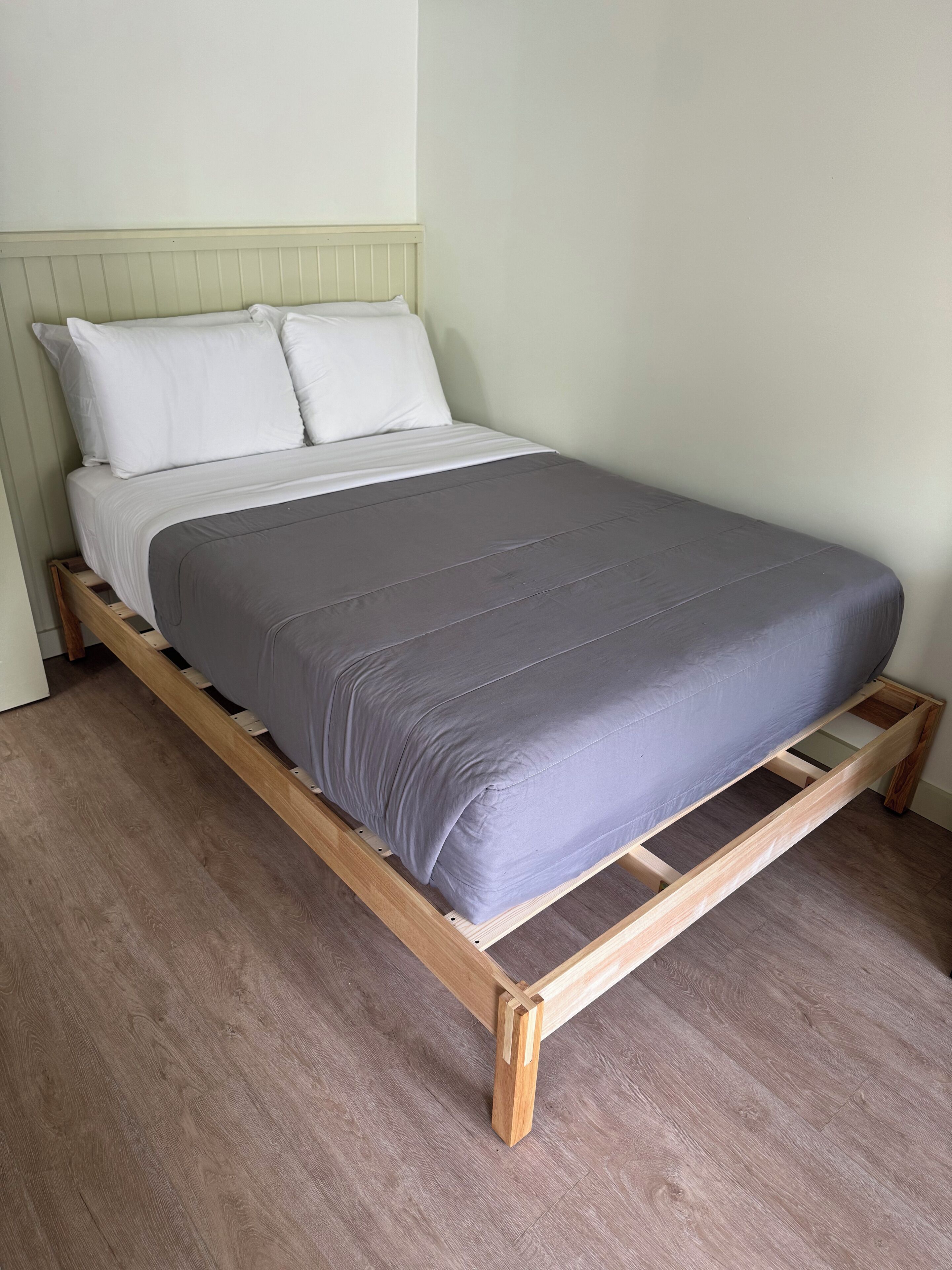 A single bed with a wooden frame and a grey mattress in a room with light green walls and wooden flooring.