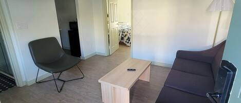 A compact living space with a sofa, a chair, a coffee table, a TV mounted on the wall, and a door leading to another room.