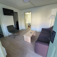 Un espacio habitable compacto con un sofá, una silla, una mesa baja, un televisor montado en la pared y una puerta que da a otra habitación.