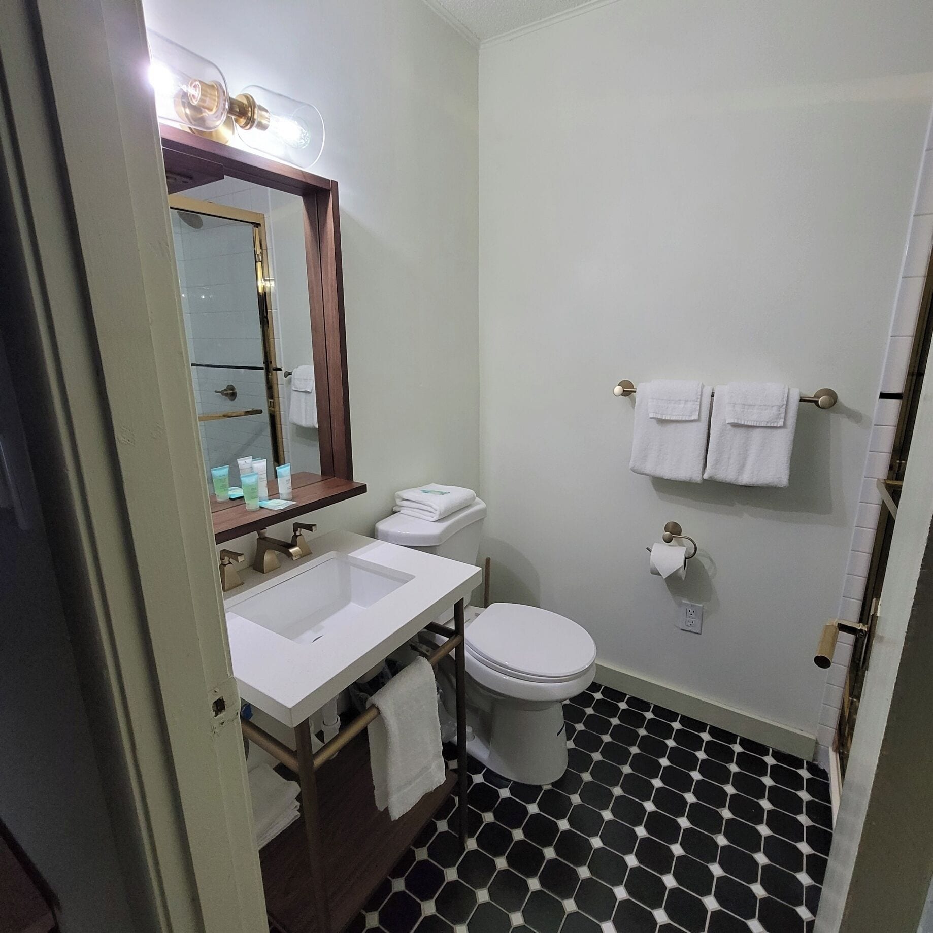 Un baño con lavamanos, inodoro y espejo blancos. Hay toallas colgadas en la pared y una puerta de ducha.