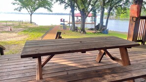 A wooden picnic table on a deck with a view of a lake and a covered area in the background. - Lakefront Gem for Family/Fishermen Getaway - 1 Hour from Dallas (Bonham)