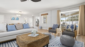 Un salon moderne comprenant un canapé, un pouf et un fauteuil, un ventilateur de plafond et une grande fenêtre avec des rideaux.