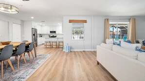 Une cuisine moderne avec des meubles blancs, un coin repas avec une table et des chaises en bois, et un salon avec un canapé et une fenêtre équipée de stores.