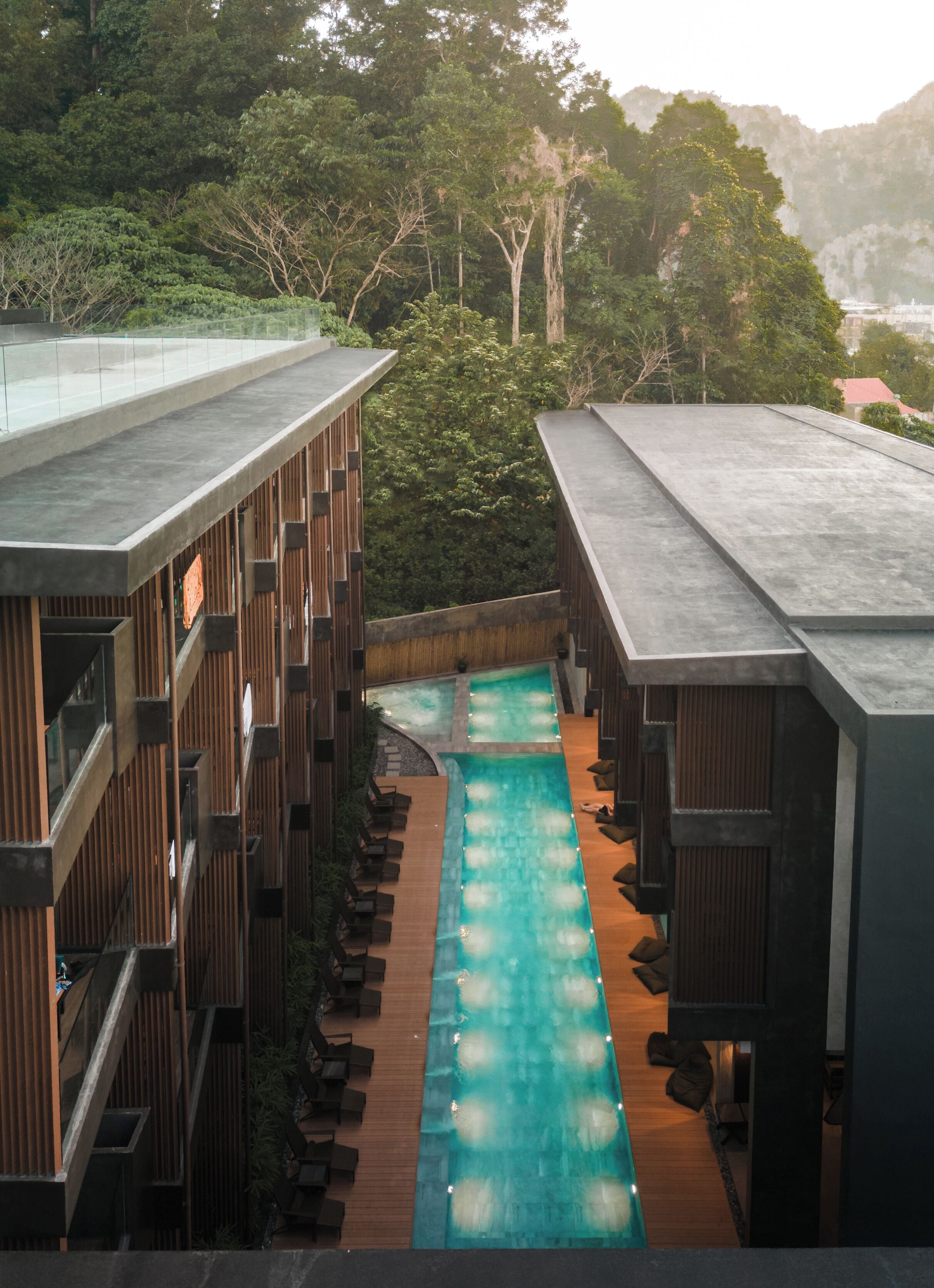 A rooftop pool area with wooden deck and lounge chairs, surrounded by dense forest and mountains.