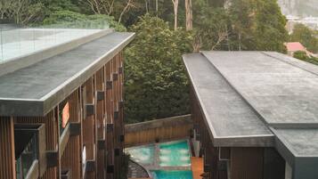 A rooftop pool area with wooden deck and lounge chairs, surrounded by dense forest and mountains.