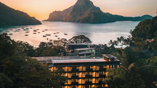 Un hotel costero de varios pisos, rodeado de exuberante vegetación y vistas al mar con barcos.