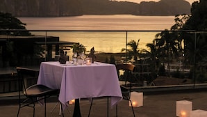 Una mesa con mantel blanco, dos sillas y velas en un balcón con vista a un cuerpo de agua y montañas al fondo.