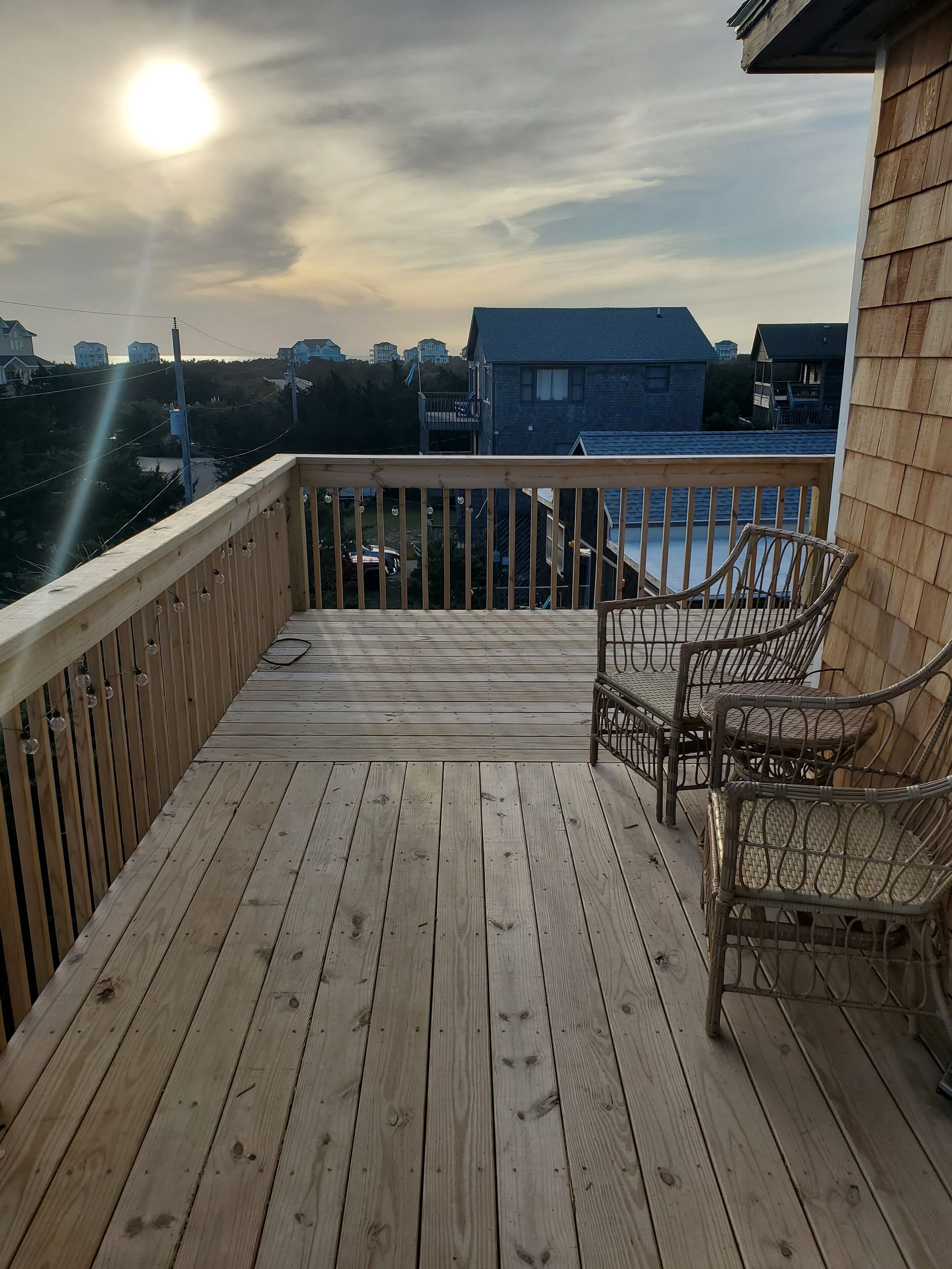Una terraza de madera con barandilla, dos sillas de mimbre y vista a casas y un cuerpo de agua al fondo.