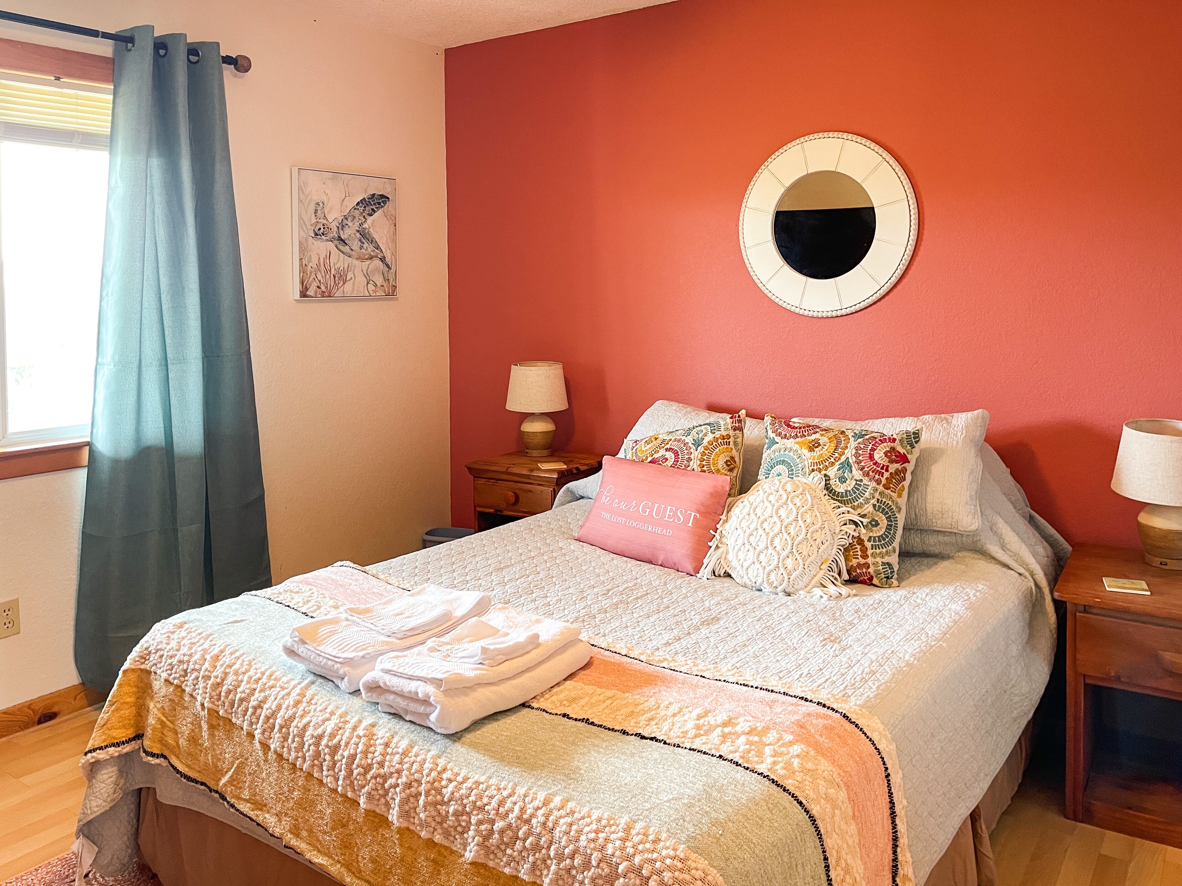 Un lit bien fait, agrémenté d’un coussin rose, d’un coussin coloré et d’un coussin blanc. On y trouve une petite table de chevet avec une lampe et une horloge murale.