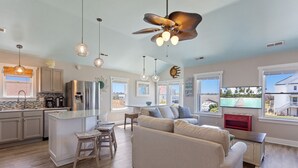 A modern kitchen with a central island, a dining area with a sofa and chairs, and a living room with a large window. - 6138 Island Life Cottage * 1 Mile to Beach (Nags Head)