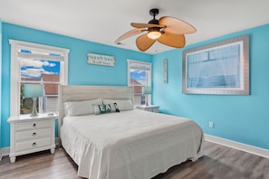 A bedroom with a bed, a ceiling fan, a window with blinds, a nightstand, and a wall-mounted picture frame.