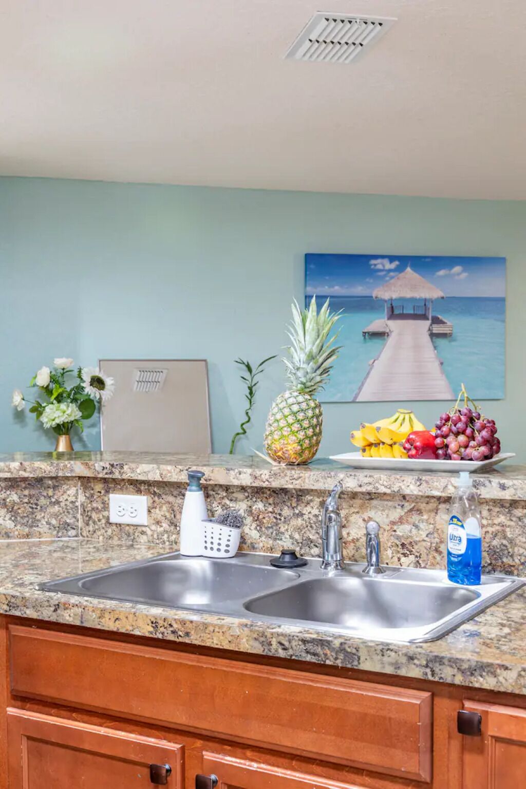 Una cocina con encimeras de granito, un lavamanos doble y un cuadro de una escena tropical.