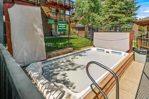 Vasca idromassaggio con terrazza in legno e ringhiere metalliche, circondata da un cortile recintato con un cartello che indica gli orari della spa.