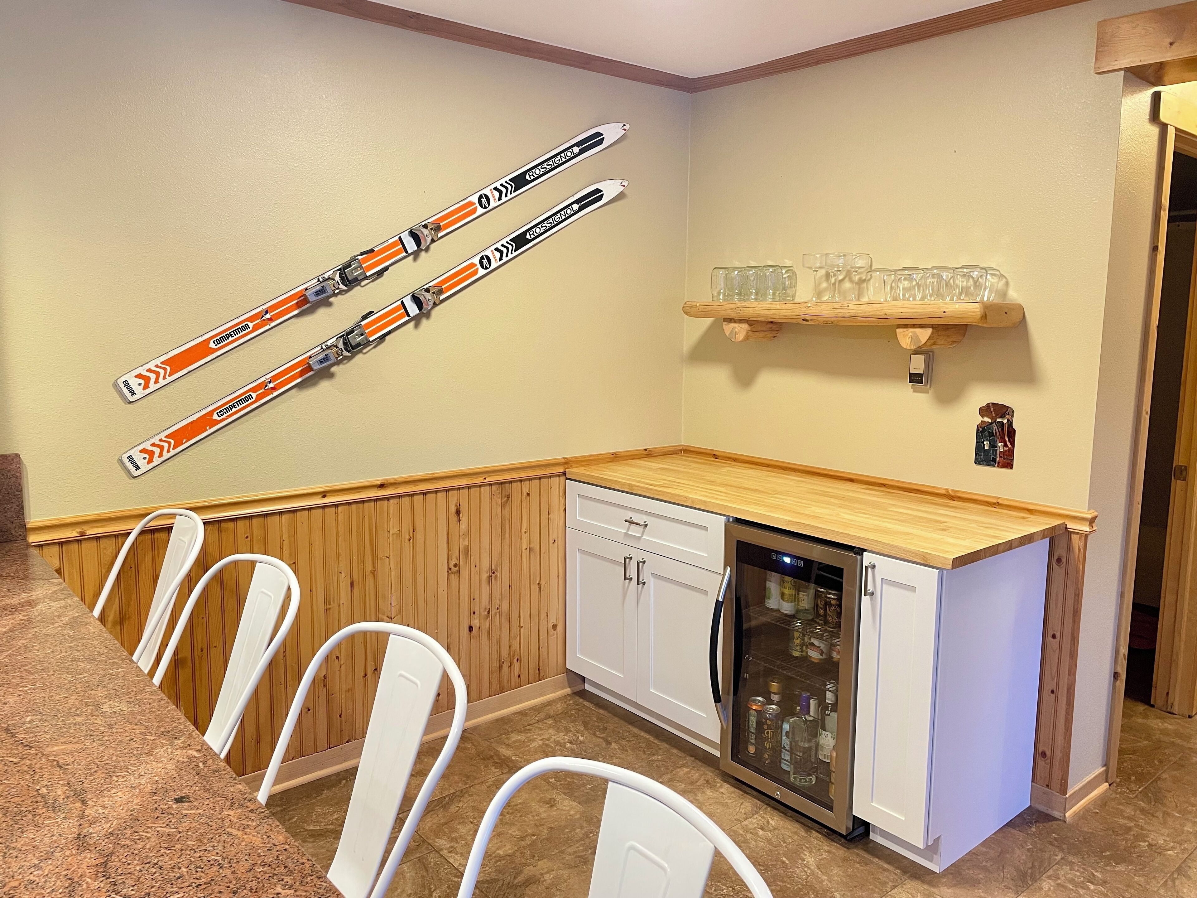 A kitchen with white cabinets, a dishwasher, and a wooden countertop. There are skis mounted on the wall and a shelf with glasses.