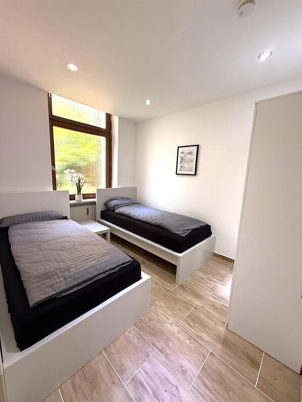 A compact bedroom with two beds, a window with a view, a framed picture on the wall, and a door.