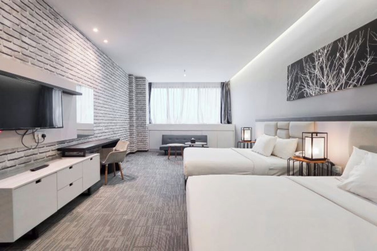A modern hotel room with a brick-patterned wall, a flat-screen TV, a desk with a chair, two beds, and a large window with curtains.