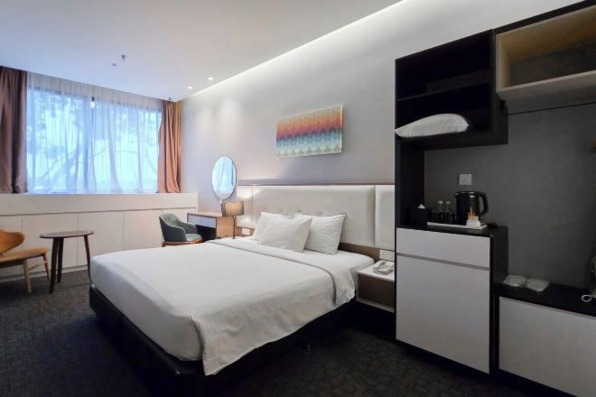 A modern hotel room with a large bed, a desk, a chair, a small table, a coffee machine, and a painting on the wall.