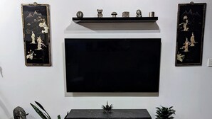 Una sala de estar con una mesa negra, un tablero de ajedrez, un televisor montado en la pared y objetos decorativos en estantes.