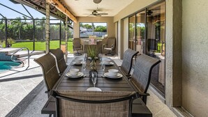 Un patio exterior con mesa de comedor preparada para cuatro personas, rodeado de sillas, una piscina y vistas a una casa y vegetación.