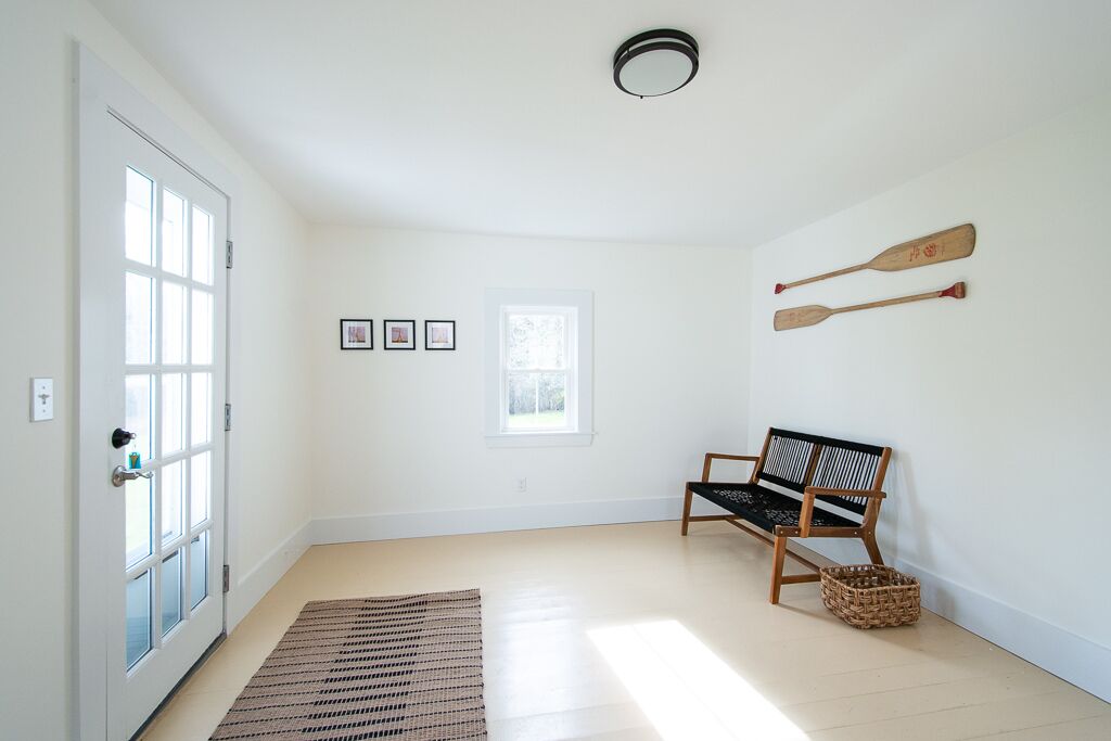Un pasillo luminoso con un banco de madera, un tapiz tejido y dos palas de madera en la pared.