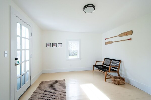 Un pasillo luminoso con un banco de madera, un tapiz tejido y dos palas de madera en la pared.