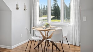 A dining area with a round table, white chairs, a bowl of fruit, and a view of a forest through large windows. - Perfect Retreat for 2 | Studio Above Bakery, Dogs! (Sister Bay)