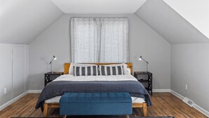 A bedroom with a sloped ceiling, a bed with a wooden headboard, two bedside tables with lamps, a window with curtains, and a bench at the foot of the bed. - Perfect Retreat for 2 | Studio Above Bakery, Dogs! (Sister Bay)