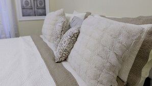 Una cama con colcha blanca y beige, almohadas estampadas y una ventana con diseño decorativo.