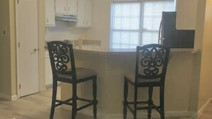 Un espacio habitable compacto con una zona de comedor que incluye dos sillas y una mesa con cubierta de vidrio. Se ve una kitchenette al fondo, y una puerta da a otra habitación.