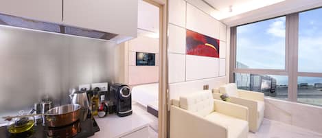 A modern hotel room with a kitchen area, a large window with a view, and a seating area with a sofa and armchairs.