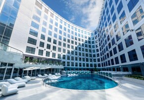 A modern hotel with a large indoor swimming pool, sun loungers, and a curved glass facade.