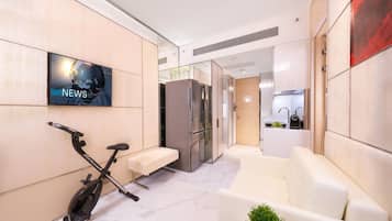 A modern living area with a TV displaying 'NEWS', a stationary bike, a sofa, and a kitchen in the background.