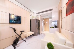 A modern living area with a TV displaying 'NEWS', a stationary bike, a sofa, and a kitchen in the background.