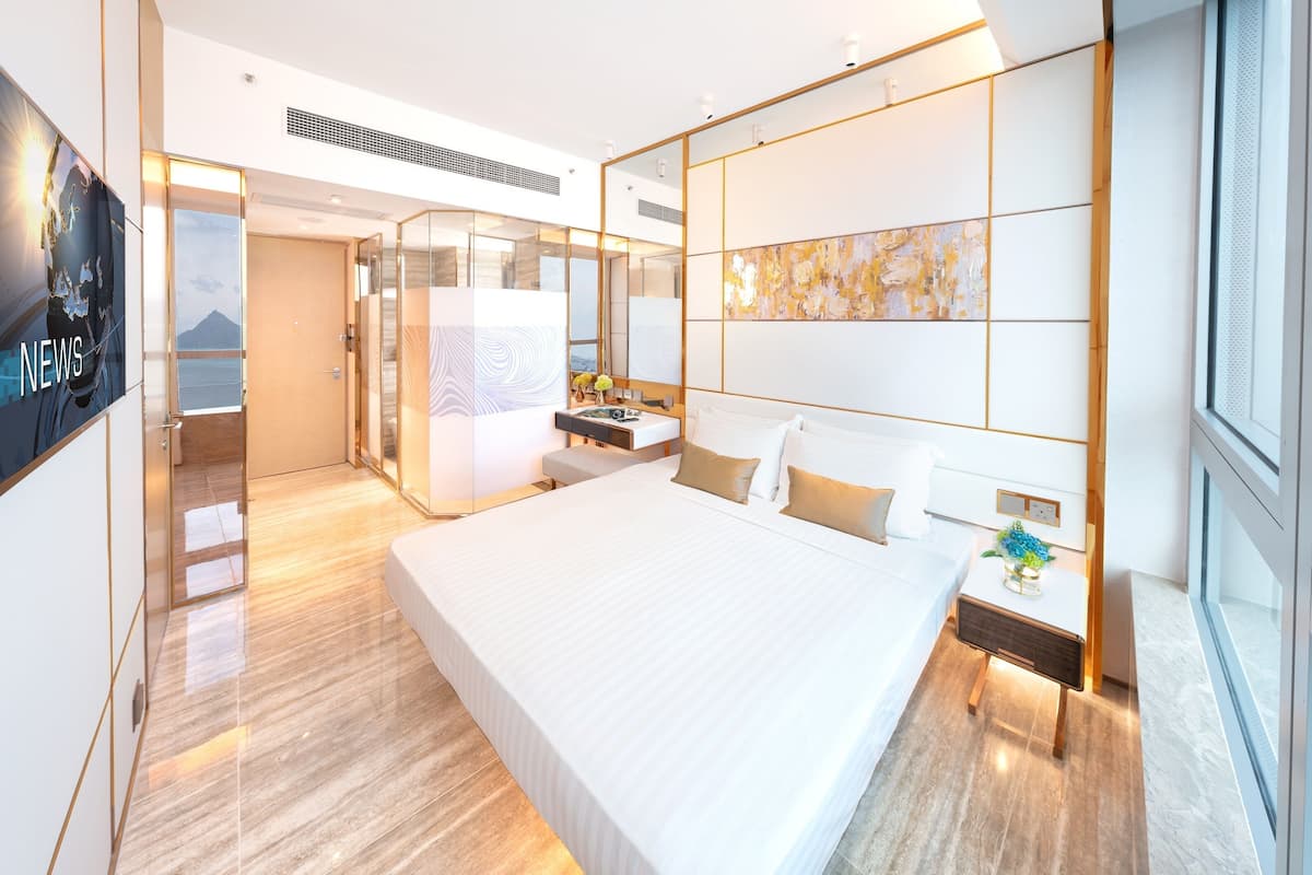 Ein modernes Hotelzimmer mit einem großen Bett, einem Nachttisch mit Lampe, einem Spiegel und Meerblick durch eine Glastür.