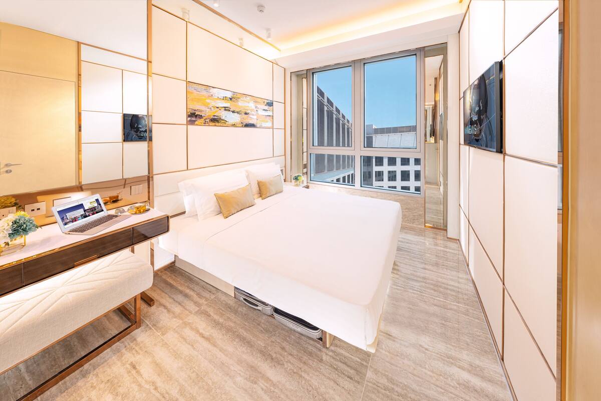 Ein modernes Hotelzimmer mit einem großen Bett, einem Schreibtisch mit einem Laptop und Blick auf die Stadt durch große Fenster.
