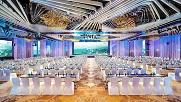 A conference room with rows of white chairs and tables, a large screen at the front, and a modern ceiling design.