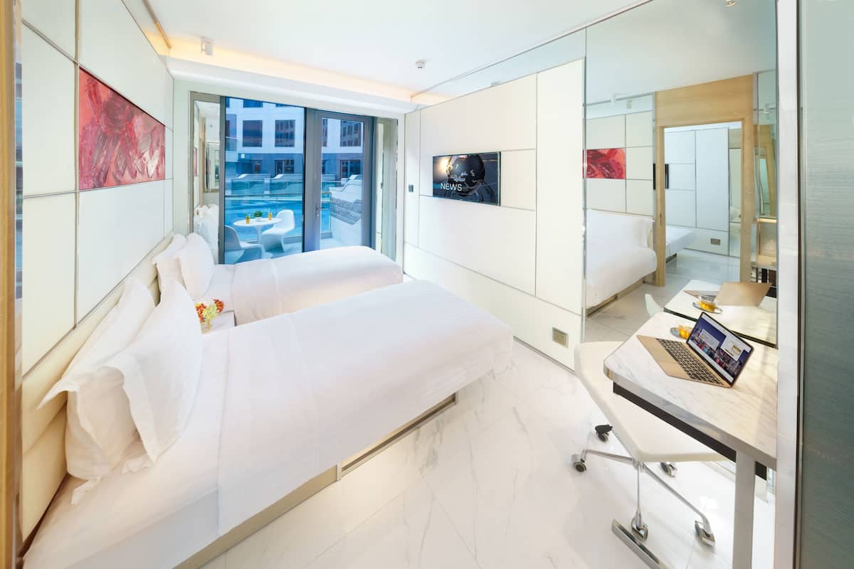 Ein modernes Hotelzimmer mit Bett, Schreibtisch mit Laptop, Fernseher und großem Fenster mit Blick auf einen Poolbereich.