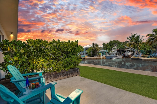 A waterfront patio with a view of the sunset, a pool, and lounge chairs.