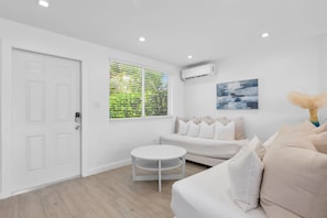 Un salon moderne avec un canapé blanc, une table basse ronde et une fenêtre donnant sur un feuillage verdoyant.