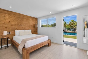 A bedroom with a wooden headboard, a bed with white bedding, a bedside lamp, a window with blinds, and a view of a pool and palm trees. - 2. Waterfront Key Largo Studio / Fish & Relax (Key Largo)